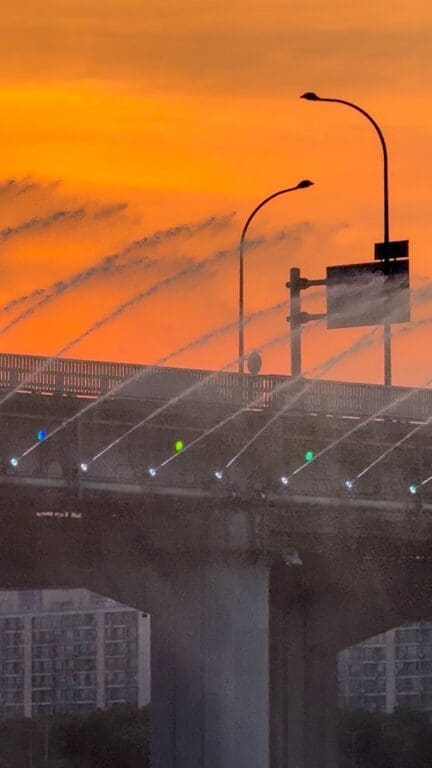 Close-up shot of the Banpo Bridge water display nozzles spraying mist against a bright orange sunset sky with street lamp posts in Seoul.