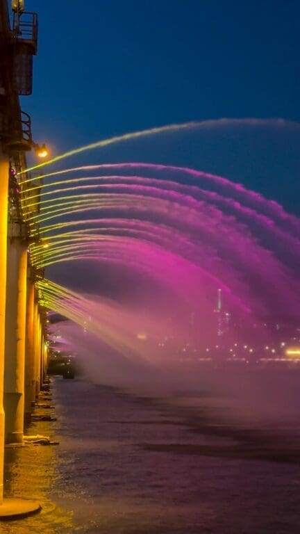 Low-angle night view of the pink and purple illuminated Banpo Bridge fountain streams shooting over the water.