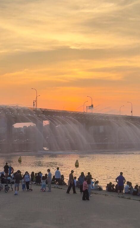 Crowds of people sitting along the Han River bank at dusk watching the Banpo Bridge Rainbow fountain spray water into the river against a soft orange sunset sky in Seoul.