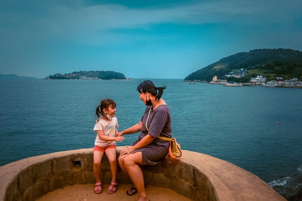 Maemiseong (매미성) | How to Visit Geoje's Magical Castle 6 mom and daughter sitting on Maemiseong 메미성 with sea behind them