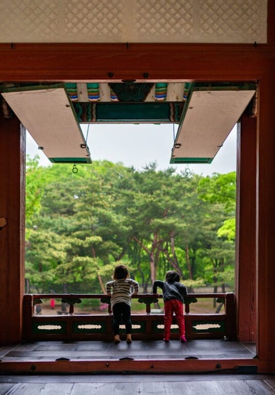 Two young children with backpacks leaning against a dark wooden railing, looking out of an open traditional Korean window frame onto a lush green forest.