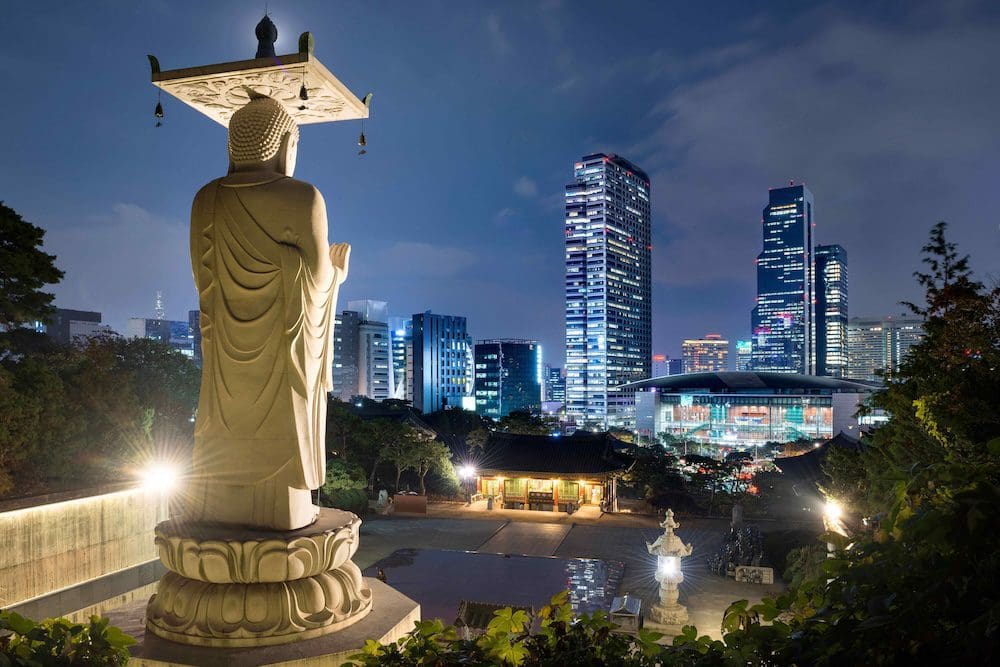 The 23-meter stone Maitreya Buddha statue at Bongeunsa Temple illuminated at night, looking out over the futuristic glass skyscrapers of Starfield Coex Mall in Gangnam Seoul.